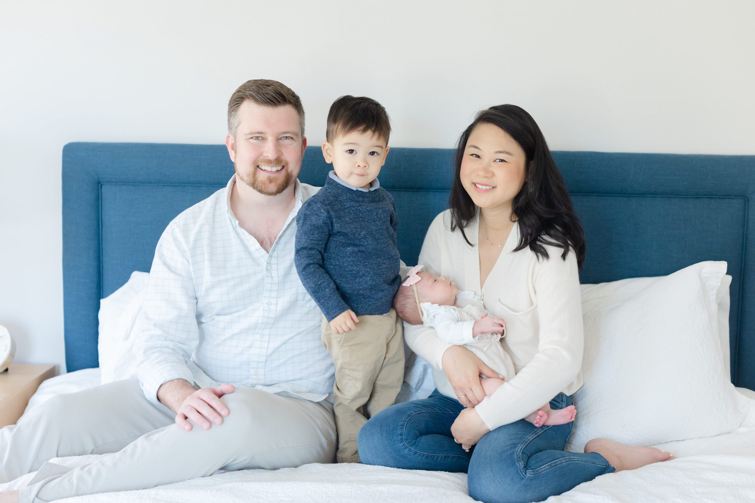 A family of four including a mom, dad, brother and baby sister smile while sitting on a bed in Westfield NJ.